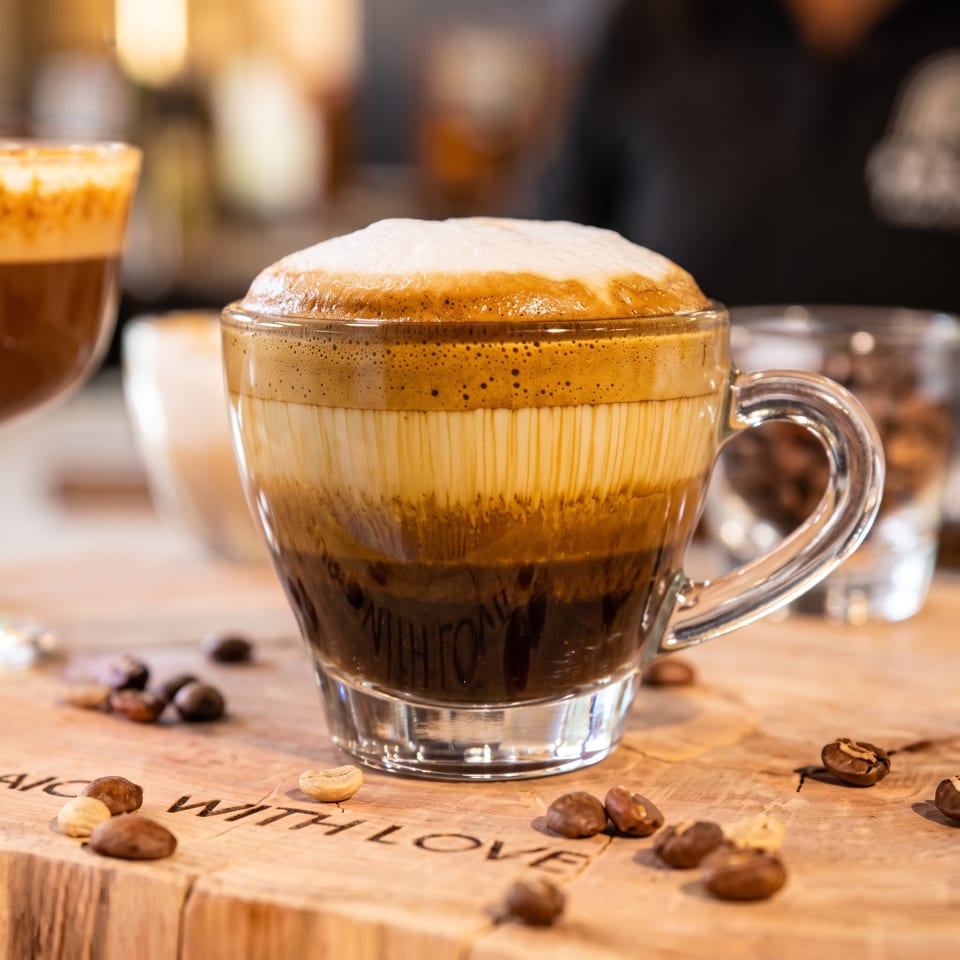 Layered coffee with creamy foam in a glass cup, surrounded by coffee beans on a wooden café counter.