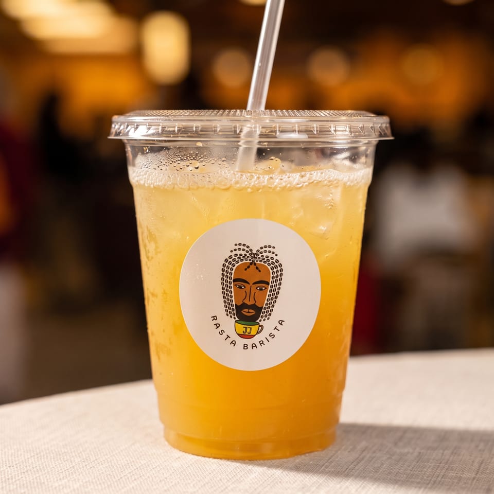 Bubbly golden iced drink in a lidded cup with straw and Rasta Barista logo, resting on a table.