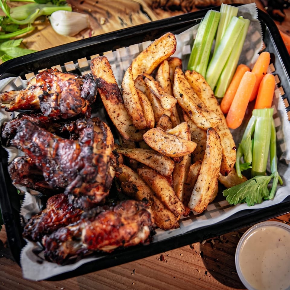 Basket of grilled chicken wings, seasoned potato wedges, carrot and celery sticks, and dipping sauce on a wooden table