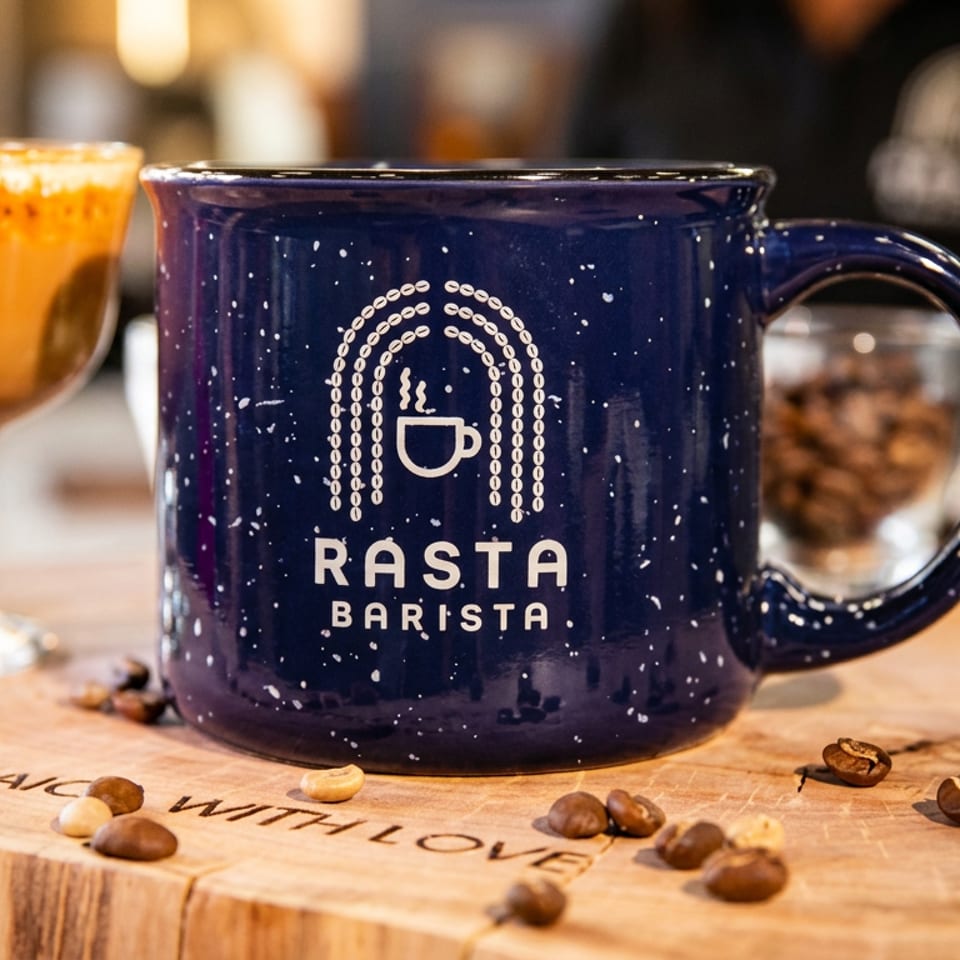 A Rasta Barista mug and a coffee drink in a glass on a wooden slab with coffee beans.